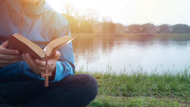 Jugendliche Person sitzt mit aufgeschlagener Bibel vor Gewässer